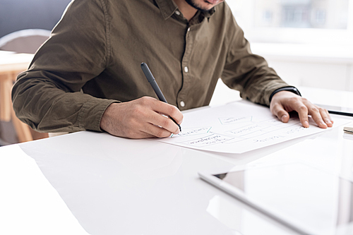 Young busy economist drawing flow chart on paper while sitting by desk in office and preparing report