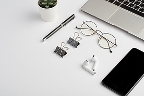 Wireless earphones, eyeglasses, pen, clips, mobile phone and laptop keypad on white desk that is workplace of employee