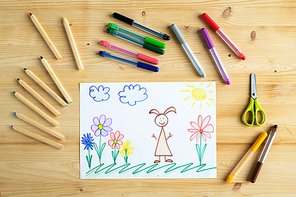 Drawing of child or schoolkid of elementary school lying on wooden table with crayons and scissors near by