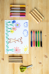 Overview of drawing of elementary schoolchild on wooden table with several sets of pencils and crayons around