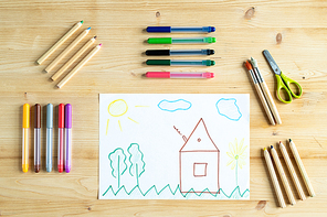 Pencils, pens, scissors and paintbrushes surrounding kid drawing of house outdoors on wooden table