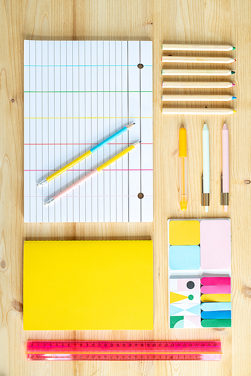 Wooden table background with sets of crayons, pens and pencils, erasers, ruler, book and lined sheet of paper