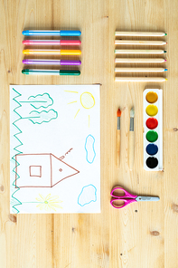 Child drawing on sheet of paper surrounded by sets of crayons and highlighters, scissors, paintbrushes and watercolor paints