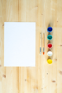 Blank sheet of paper, two paintbrushes and row of colorful paints or gouaches on wooden background