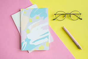 Top view of eyeglasses and pen on yellow background and two notebooks on pink background