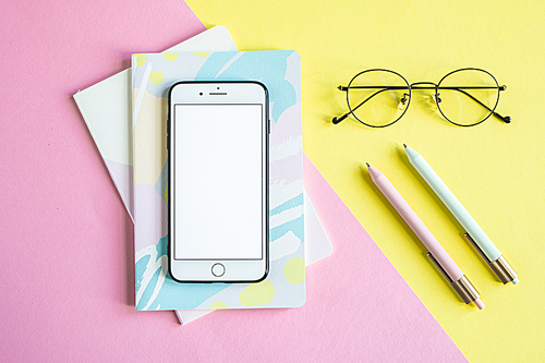 Smartphone on two notebooks over pink background and eyeglasses and pens on yellow background