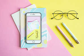 Smartphone with image of its screen, notebooks, pens and eyeglasses over pink and yellow background