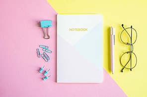 Several pins, clip, notebook, pen and eyeglasses on pink and yellow background