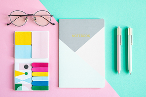 Overview of notebook, two pens, eyeglasses and set of office supplies on pink and blue background