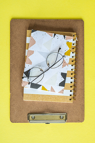 Eyeglasses on top of notepad, notebook and clipboard over yellow background in isolation