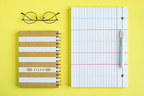 Eyeglasses, closed notebook with spiral binder, pen and lined paper on yellow background