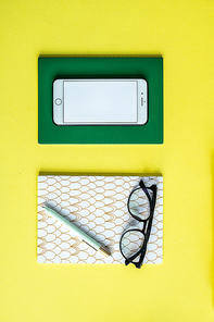 Smartphone on green notebook and pen with eyeglasses on copybook over yellow background