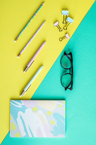 Overview of pens and clips on yellow background and notebook with eyeglasses on blue background
