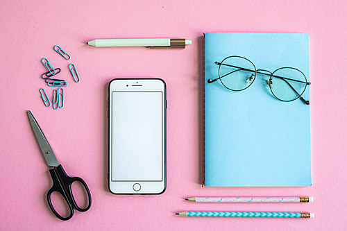 Smartphone, scissors, notebook with eyeglasses, clips, pen and pencils on pink background