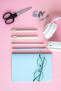 Pens, pencils, clips, scissors, headphones, eyeglasses and notebook in blue cover on pink background