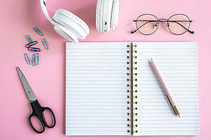 Open copybook with pen, eyeglasses, scissors, clips and headphones on pink background