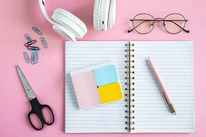 Open notebook, erasers, pen, clips, scissors, eyeglasses and headphones on pink background