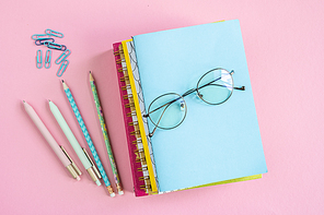 Stack of notebooks with eyeglasses on top with groups of clips, pens and pencils near by on pink background