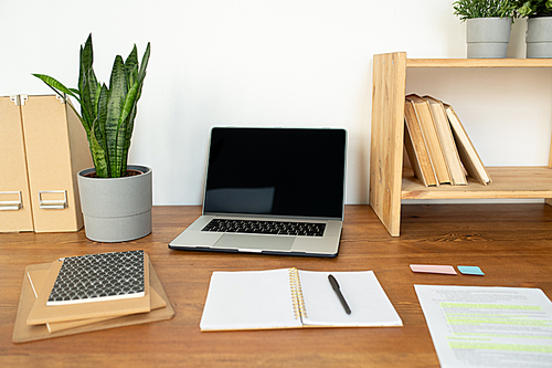 Wooden shelf with books, open copybook with pen, laptop, potted plant and stack of notepads on desk