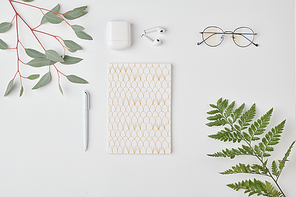 Flatlay of notebook with pen, eyeglasses and earphones on white background surrounded by green plants