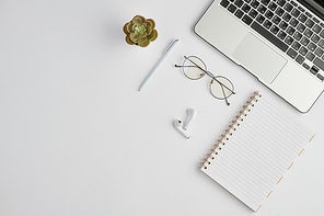 Wireless earphones, eyeglasses, pen, notebook, small domestic plant and laptop keypad on white desk
