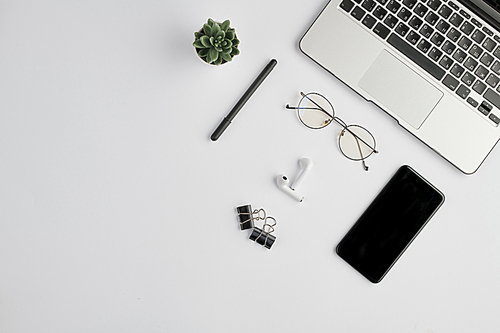 Wireless earphones, eyeglasses, pen, clips, smartphone, small domestic plant and laptop keypad on white desk