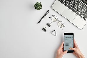 Hands of female employee holding smartphone with monthly planner on screen while organizing work by desk