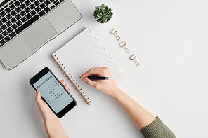 Hands of female employee holding smartphone with monthly planner on screen and pen over blank page of notebook