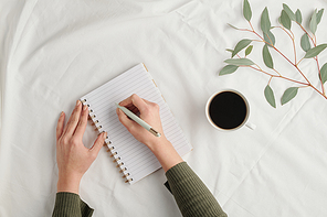 Overview of hands of young businesswoman with pen making notes in notebook while having cup of coffee