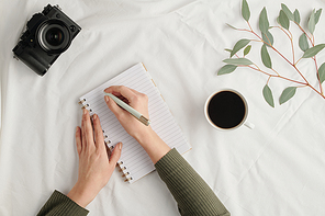 Hands of young woman with pen over open notebook surrounded by photocamera, cup of coffee and branch with green leaves