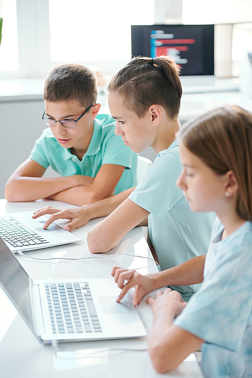 Row of serious cute schoolkids in casualwear sitting by desk in front of laptops while preparing for school seminar at break