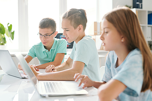 Three young schoolkids in casualwear sitting in computer classroom while concentrating on network at lesson