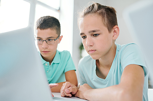 Two clever schoolboys sitting by desk in front of laptop display while searching for online data for seminar or project