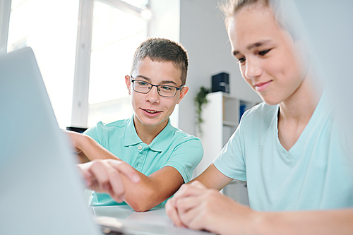 Cute schoolboy and his classmate looking at laptop screen while sitting in classroom and surfing in the net