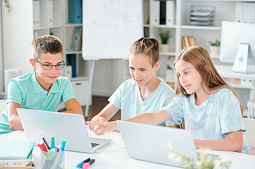 Three schoolmates pointing at something curious on laptop display while discussing it in classroom