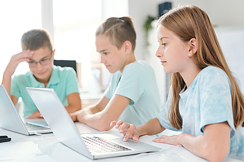 Young serious schoolgirl and her classmates working individually in front of laptops while sitting by desk