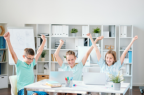 Three ecstatic successful schoolmates keeping their hands raised while expressing joy in front of laptops