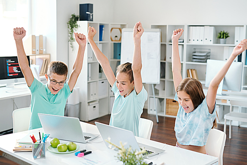 Cheerful middle school students with raised hands looking at laptop displays while expressing excitement