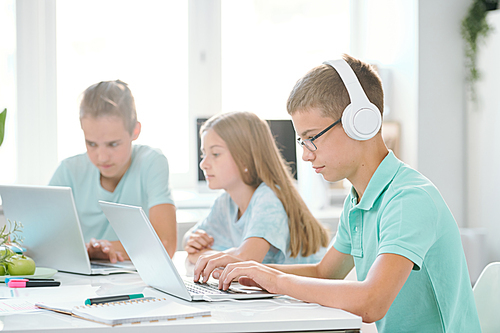 One of youthful classmates in headphones looking at laptop display while watching online video by desk