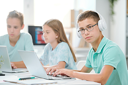 Handsome schoolboy in headphones looking at you while typing on laptop keypad by desk with classmates on background