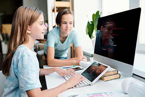 Pretty youthful girl typing on computer desktop keypad while looking at monitor and preparing home assignment