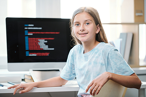 Attractive middle school student in casualwear looking at you while sitting by desk in front of computer monitor