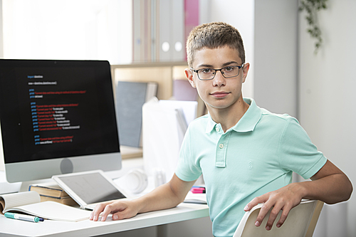 Clever boy from middle school looking at you while sitting by desk during network or homework preparation