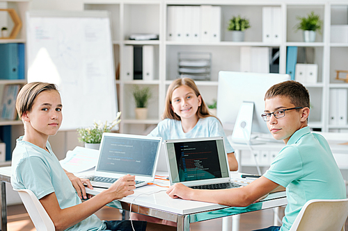 Group of clever middle school students looking at you in classroom while sitting by desk in front of laptops