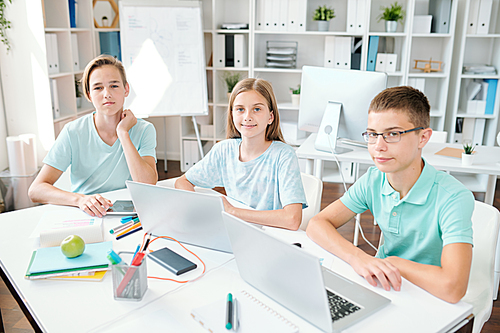 Three diligent learners of middle school looking at you while sitting by desk in classroom and doing homework