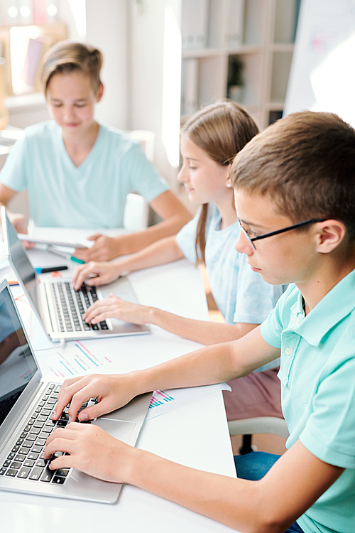 Two boys and girl in casualwear sitting by desk in front of laptops while carrying out final exam test in classroom