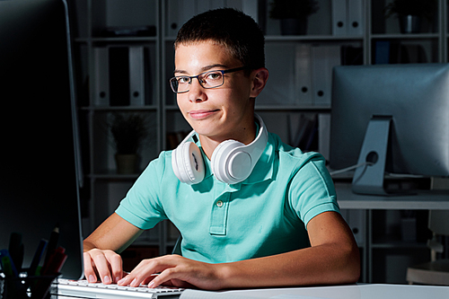 Young serious schoolboy in eyeglasses looking at you while networking late in dark classroom at school