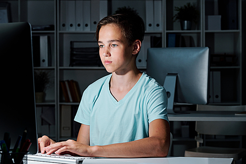 Diligent schoolboy sitting in dark classroom by desk while preparing for seminar or school project for next day