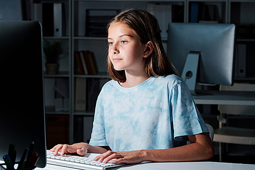 Pretty schoolgirl in t-shirt looking at computer screen while networking in the dark classroom after lessons