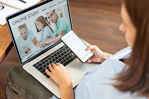 Young woman with smartphone sitting in front of laptop display while surfing on educational website